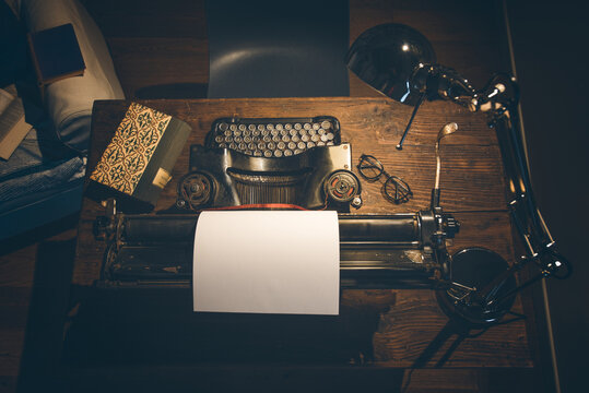 An old classic typewriter in a vintage style studio with a wooden desk lit by a lamp, wit old books all around. A moody, dark atmosphere for a writer working at night.