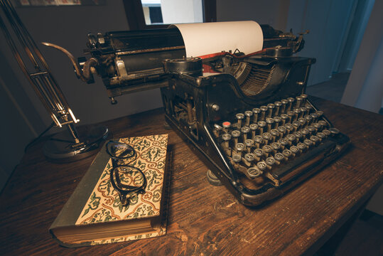 An old classic typewriter in a vintage style studio with a wooden desk lit by a lamp, wit old books all around. A moody, dark atmosphere for a writer working at night.