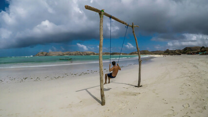 Obraz na płótnie Canvas A man swinging on a swing placed on the seashore of Tanjung Aan Beach, Lombok, Indonesia. The swing has simple wood construction. Waves wash the pillars of it. In the back there are few boats.