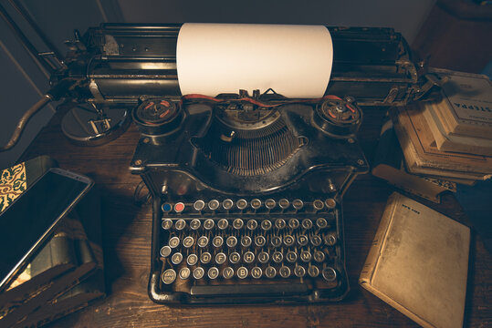 An old classic typewriter in a vintage style studio with a wooden desk lit by a lamp, wit old books all around. A moody, dark atmosphere for a writer working at night.