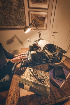 Typing hands of a writer looking for inspiration to start a new novel on his vintage typewriter in a retro style studio: a wooden desk lit by a lamp, old books and a pair of glasses.