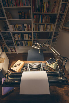 An old classic typewriter in a vintage style studio with a wooden desk lit by a lamp, wit old books all around. A moody, dark atmosphere for a writer working at night.