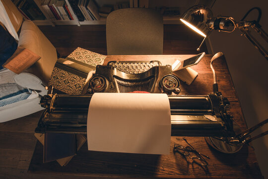 An old classic typewriter in a vintage style studio with a wooden desk lit by a lamp, wit old books all around. A moody, dark atmosphere for a writer working at night.