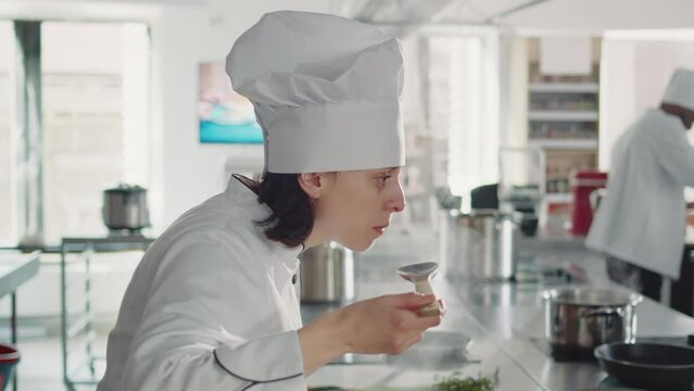 Female chef tasting liquid sauce on stove to make cuisine dish, cooking culinary recipe with fresh ingredients. Woman with uniform working in restaurant kitchen to create professional menu. Close up.
