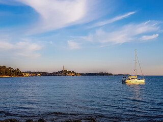 Golden hour in the harbor next to Rovinj, Croatia. The city is located in the back. A small yacht anchored to a buoy. Soft clouds on the sky. Sea water is very calm.