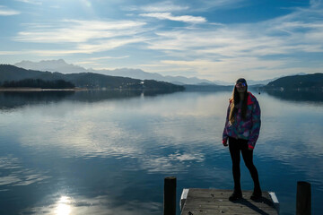 Fototapeta na wymiar Girl wearing a jacket and beanie stands at the end of a promenade on the lake. Soft reflections of the clouds in the lake. Clear but cloudy day. High mountains in the back Calmness and relaxation