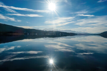 A view on a lake and Alps in the back. The calm surface of the lake is reflecting the mountains, sunbeams and clouds. Clear and sunny day. Calm and relaxed feeling.