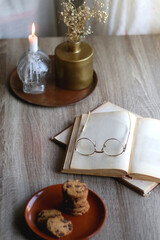 Stack of chocolate chip cookies, open book and reading glasses, lit candle and vase with flowers on the table. Hygge at home. Selective focus.