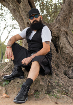 full length image bearded man wearing sunglasses, cap and skirt sitting on a base of a tree