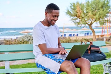 Young smiling male freelancer relaxing on beach, sitting on bench using laptop