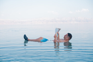 Man lies in blue water of Dead Sea. Person with newspaper in hands. Very salty water push out....