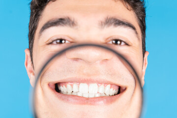 Hispanic teenager boy showing white teeth through loupe isolated on blue background. Dentistry...