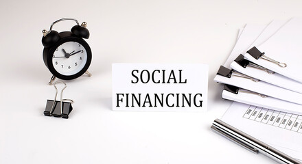 Card with text SOCIAL FINANCING on a white background, near office supplies and alarm clock. Business concept.
