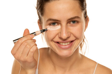 Young smiling woman applyes concealer under her eyes on white background