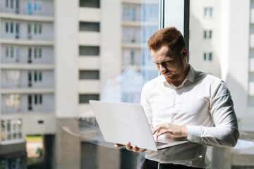 Medium shot portrait of focused young man working typing on laptop holding with one hand standing in modern office room near window, in sunny summer day on background of buildings.