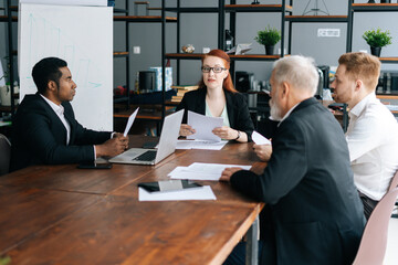 Serious female team leader in glasses holding corporate meeting with multiethnic colleagues in boardroom sitting at desk. Young businesswoman discussing project with business partners in office.
