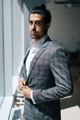 Vertical portrait of stylish handsome business man wearing fashion suit standing near window in modern office room, looking at camera. Front view of bearded businessman posing at workplace.
