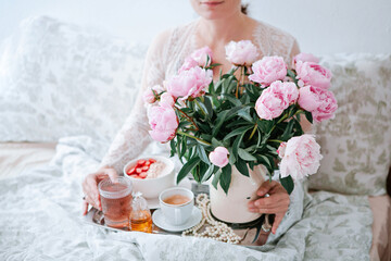 Young woman holding tray with bouquet of pink peonies flowers and a cup of black coffee and Breakfast in bed, summer morning concept. Healthy food