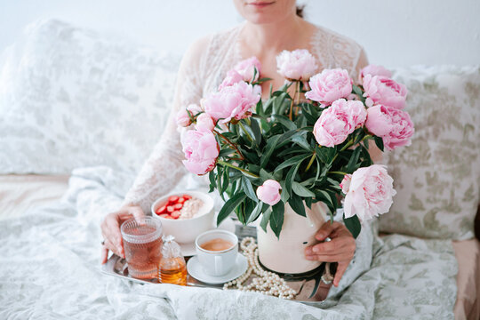 Young Woman Holding Tray With Bouquet Of Pink Peonies Flowers And A Cup Of Black Coffee And Breakfast In Bed, Summer Morning Concept. Healthy Food