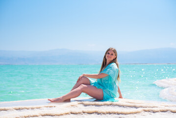 Fototapeta na wymiar Slim woman in turquoise pelerine sits on salt island of Dead Sea on background sea and mountains on horizon. Treatment and recreation in Israel, salt and mud are good for health.