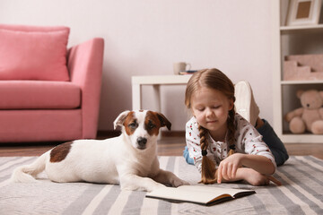 Cute little girl with her dog reading book on carpet at home. Childhood pet