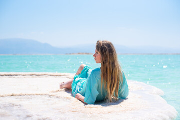 Young woman in turquoise pelerine sits on salt island of Dead Sea on background sea and mountains on horizon. Treatment and recreation in Israel, salt and mud are good for health.
