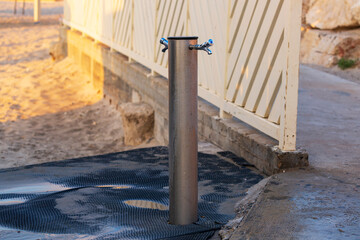 Place for washing the feet on the beach in sunny day. Outdoors water tap against the white wooden fence. Two faucets, vertical valves for drinking, washing feet and black non-slip rubber mat, pad.