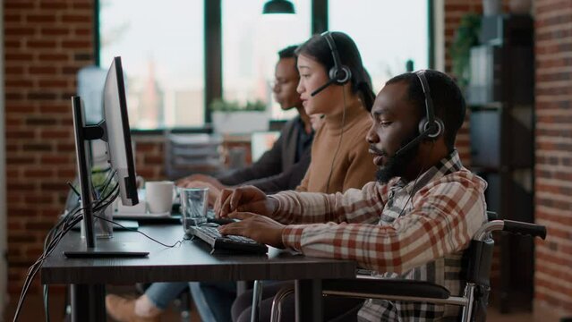 African american employee using headphones to talk to clients, helping with sales and giving telemarketing assistance at call center. Male operatortalking to people in disability friendly office.