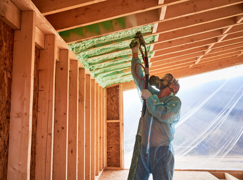 Male builder insulating wooden frame house. Man worker spraying polyurethane foam inside of future cottage, using plural component gun. Construction and insulation concept.