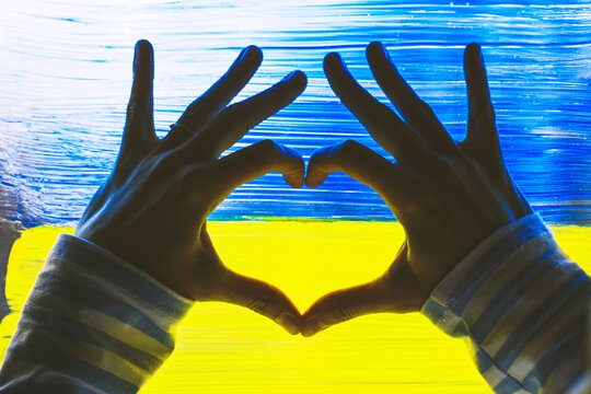 Female hands shaping heart symbol in background of yellow-blue flag of Ukraine on window. Human hands made heart shape on image of flag of Ukraine on glass. Support Ukrainians. Concepts symbol