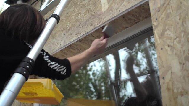 Female worker standing on ladder coats the wooden surface of outer window slope with a primer. Hand with brush
