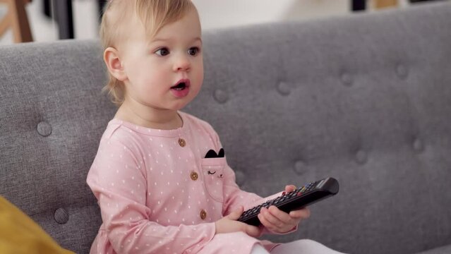 Two years old child watching tv sitting on grey carpet at home alone using tv remote.