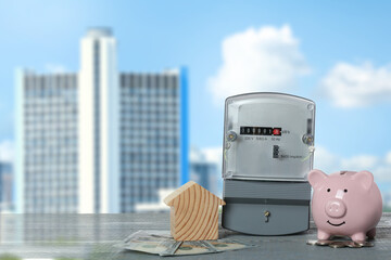 Electricity meter, house model, money and piggy bank on grey wooden table against blurred view of...