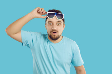 Young man surprised by something extraordinary. Happy handsome guy in blue T shirt lifts his sunglasses, looks at camera with funny shocked face expression and says WOW. Studio shot on blue background