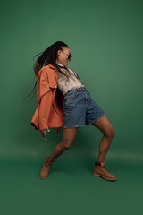 Emotional african girl in shorts and shirt dancing, having fun isolated on dark green background. Concept of beauty, art, fashion, youth and emotions