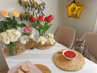 Obraz na płótnie Canvas The cat sits at a table in a white kitchen with a plate of borscht against the background of bouquets of flowers.