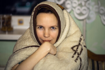 A woman covered with a blanket sits at home. So cold. Woman forty years old looking at the camera.