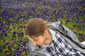 A teenage girl on a lawn with purple flowers in the park. A girl and a spring flower. An open romantic portrait of a young happy and attractive girl in a plaid hoodie enjoying a carefree pastime