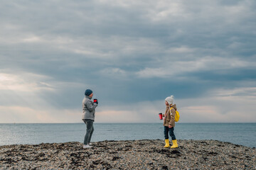 grandmother and granddaughter on a spring day off are engaged in health, walking along the seashore an adult woman and a little girl in rubber boots, with a yellow backpack