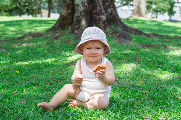 Adorable baby girl in white clothing and panama is playing with dry yellow leaves on grass. Summer sunny day in park.