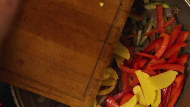 A woman's hand from the cutting board tosses chopped red bell peppers and potatoes into the cauldron where the meat and vegetables are roasting. Steam rises from the bottom of the wok