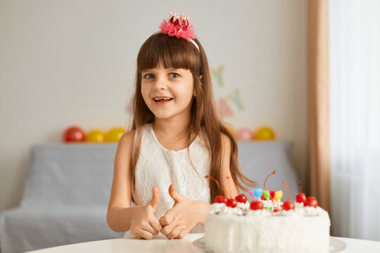 Image of cute little girl sitting at table with birthday cake at home against a backdrop of balloons, looking at camera with excited happy expression, expressing happiness.