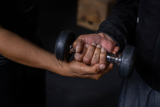 Latin American senior male with his unrecognizable personal trainer do rehabilitation and therapy exercises holding a dumbbell in the gym. Healthy lifestyle concept.