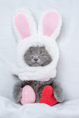 Funny kitten wearing easter rabbits ears sleeps with red heart and painted egg on a bed under warm white blanket at home. Top down view