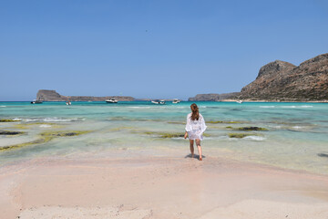 Pretty blonde girl in white beach dress walking towards turquoise blue Mediterranean sea on a hot summer day in Greece