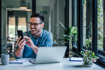 Cheerful businessman checking smart phone while using laptop at table