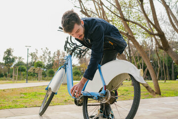 Young handsome man is checking tire on his bike on city park.