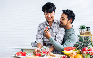 Gay LGBT sweet Asian couple wearing pajamas, smiling with happiness and love, eating, feeding watermalon, healthy fruits for breakfast in kitchen at home in the morning with sunlight. 