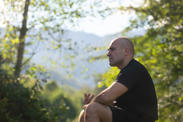 Naklejka na ściany i meble a man of strong athletic build sits in a forest in the background of a mountain, he thought about something and went deep into his thoughts