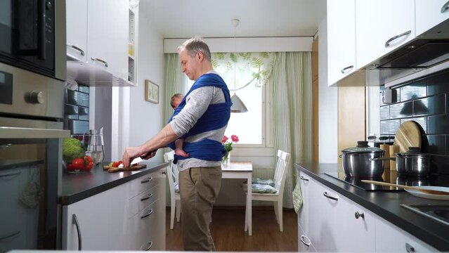 Father with little baby sleeping in sling cooking in the kitchen. Man on paternity leave working household chores. Dad enjoying fatherhood responsibility caring for child